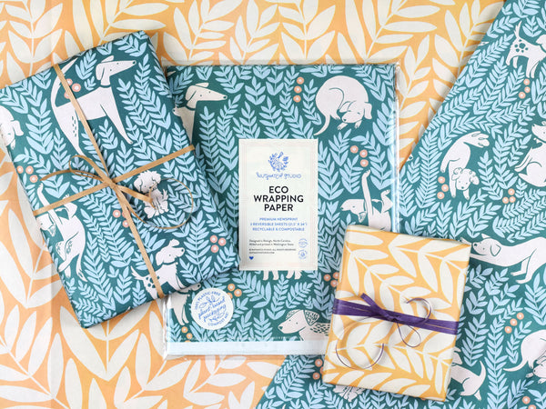 Package of the dog park wrapping paper surrounded by two wrapped gifts, each featuring a different side of the wrapping paper: one with the illustrated dogs pattern, the other featuring large illustrated leafy ferns.