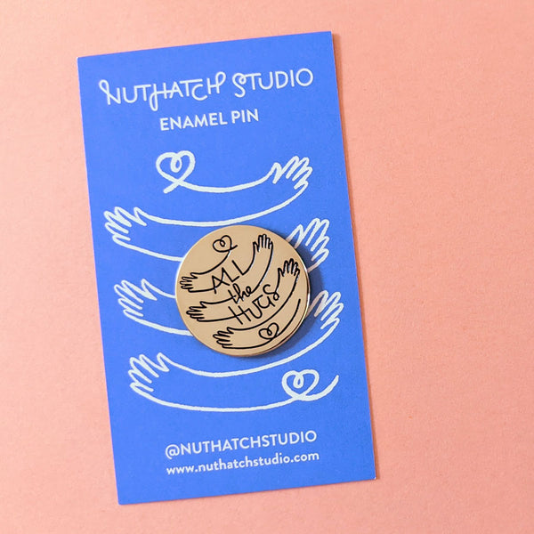 Gold enamel circle pin with a one-line illustration of the words All the Hugs connected with hearts and hands in a hugging motion.