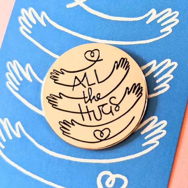 Close-up detail image of gold circle enamel pin with All the Hugs illustration on a blue backer card.