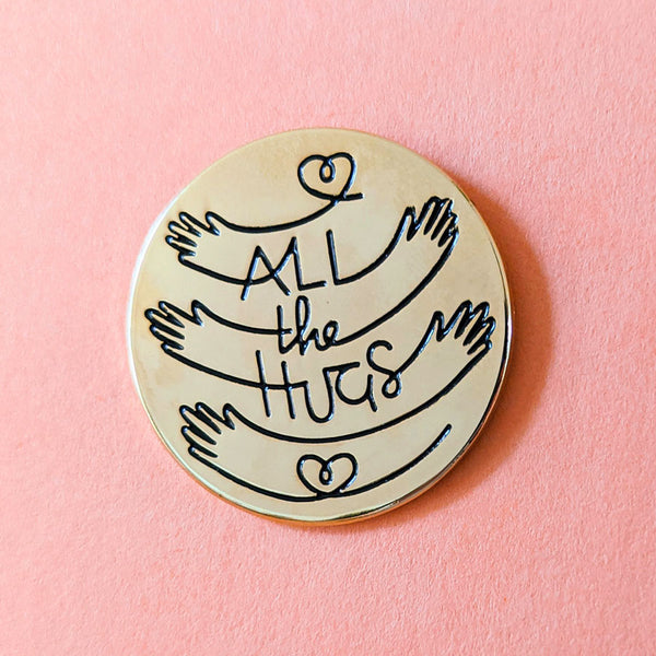 Close-up detail image of gold circle enamel pin with All the Hugs illustration on a pink background.