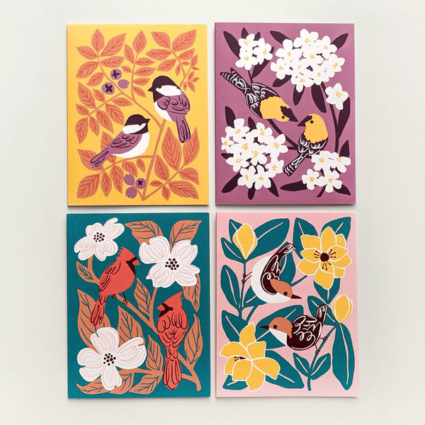 Grid of four colorful greeting cards each with a different illustrated design of two birds (chickadees, goldfinches, cardinals, and nuthatches) surrounded by leaves and flowers.