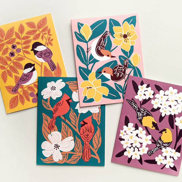 Four colorful greeting cards side by side each with a different illustrated design of two birds (chickadees, goldfinches, cardinals, and nuthatches) surrounded by leaves and flowers.