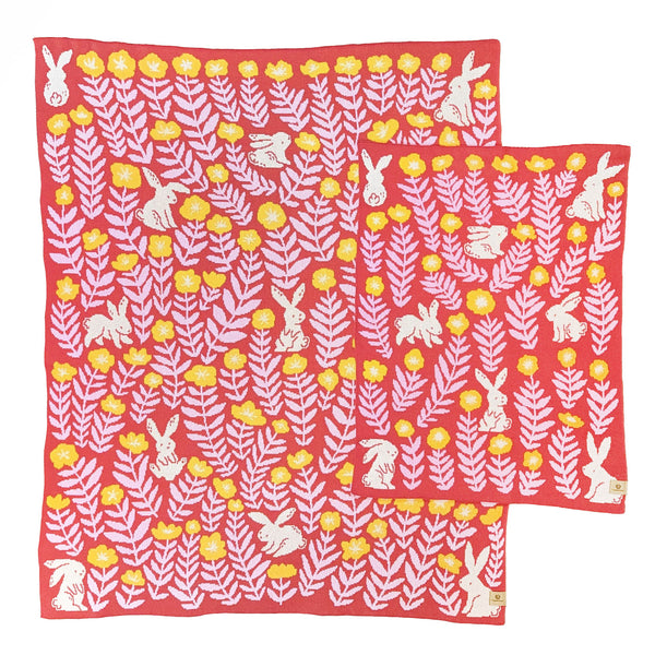 Crib and throw-size knit blankets illustrated with rabbits and flowers. The background color is a warm coral red with cream-colored bunnies, marigold yellow flowers, and soft pink leaves.