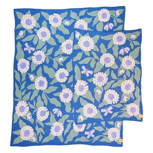 Crib and throw-size knit blankets illustrated with daisies, butterflies, and ladybugs. The background color is a soft cerulean blue with soft pink daisies, light sage green leaves, and lavender butterflies.