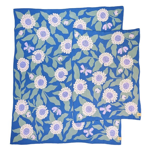 Crib and throw-size knit blankets illustrated with daisies, butterflies, and ladybugs. The background color is a soft cerulean blue with soft pink daisies, light sage green leaves, and lavender butterflies.