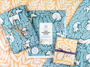 Package of the dog park wrapping paper surrounded by two wrapped gifts, each featuring a different side of the wrapping paper: one with the illustrated dogs pattern, the other featuring large illustrated leafy ferns.