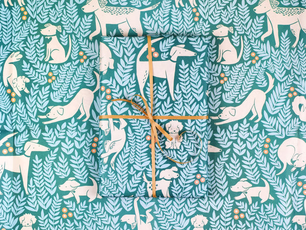 A gift wrapped in the Dog Park paper featuring cream-colored illustrated dogs in all shapes and sizes on a dark green background surrounded by light blue leafy plants and round yellow flowers.
