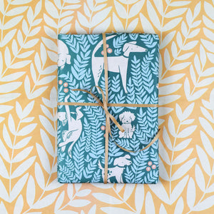 A gift wrapped in the Dog Park paper on a backdrop of the reverse side of the paper featuring yellow and cream-colored leafy ferns.