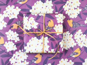 A gift wrapped in the Goldfinch paper featuring cheery yellow illustrated birds perched among bursting oleander blooms on a vibrant plum-colored backdrop.