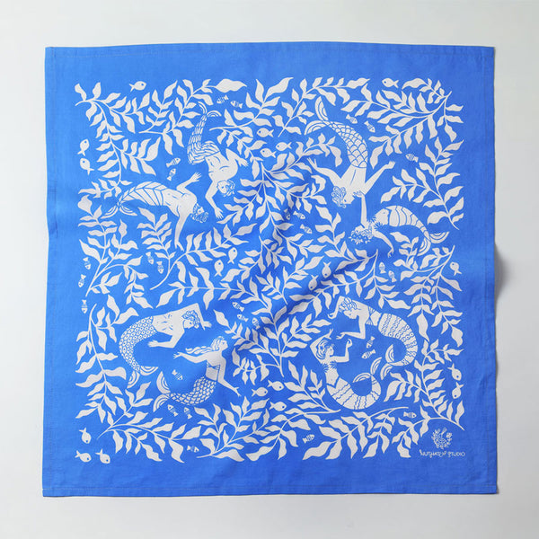 Flat view of cerulean blue and white bandana with a block printed style illustration of dancing mermaids, mermen, flowing kelp leaves, and fish.